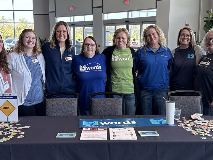 eight members of the WORDS team pose for a photo behind a table with information about the WORDS Project at the NeMTSS conference