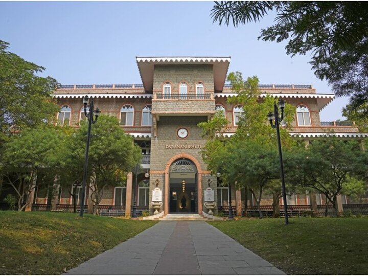 A multi-story building with a clock above the entrance, surrounded by trees and a paved walkway.
