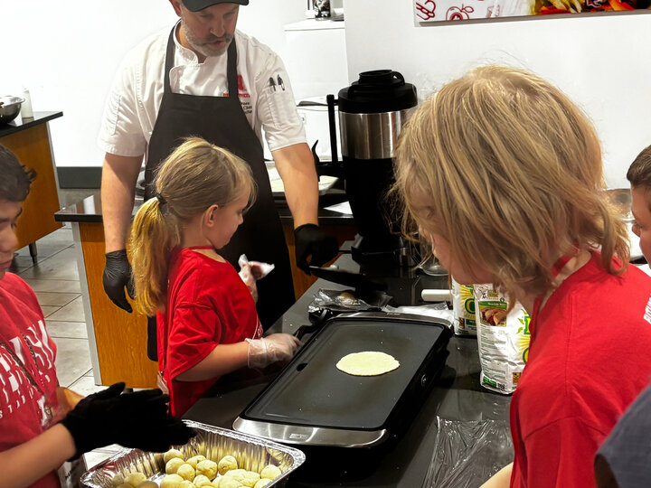 Chef guides children cooking on a griddle, surrounded by ingredients in a kitchen setting.