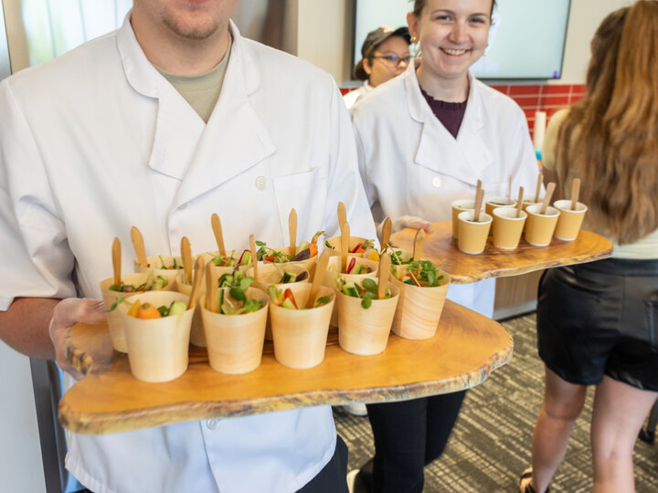 Two chefs holding trays of small wooden cups with colorful salads in a casual indoor setting.