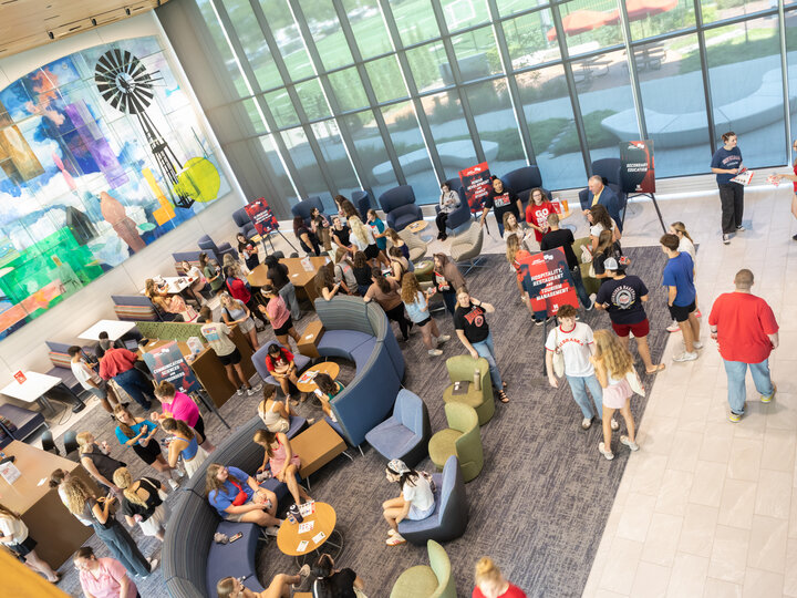 Crowded indoor event with people socializing, booths, colorful mural, and large windows.