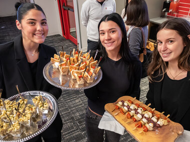 three HRTM students dressed in black shirts each hold serving trays with food samples while smiling for a group photo at the UNLimited Hospitality event