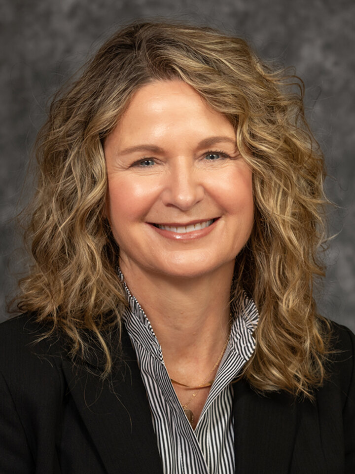 Smiling woman with curly blonde hair wearing a black suit jacket and gray striped shirt against a gray textured background.