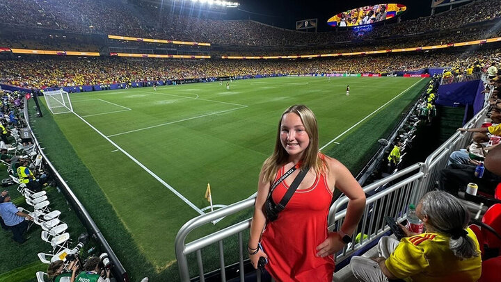 Smiling woman in a red dress stands in a stadium overlooking a soccer match with a packed crowd.
