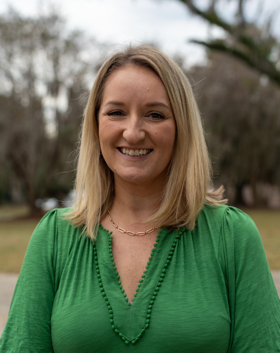 Smiling woman with blonde hair wearing a green blouse, standing outdoors with trees in the background.