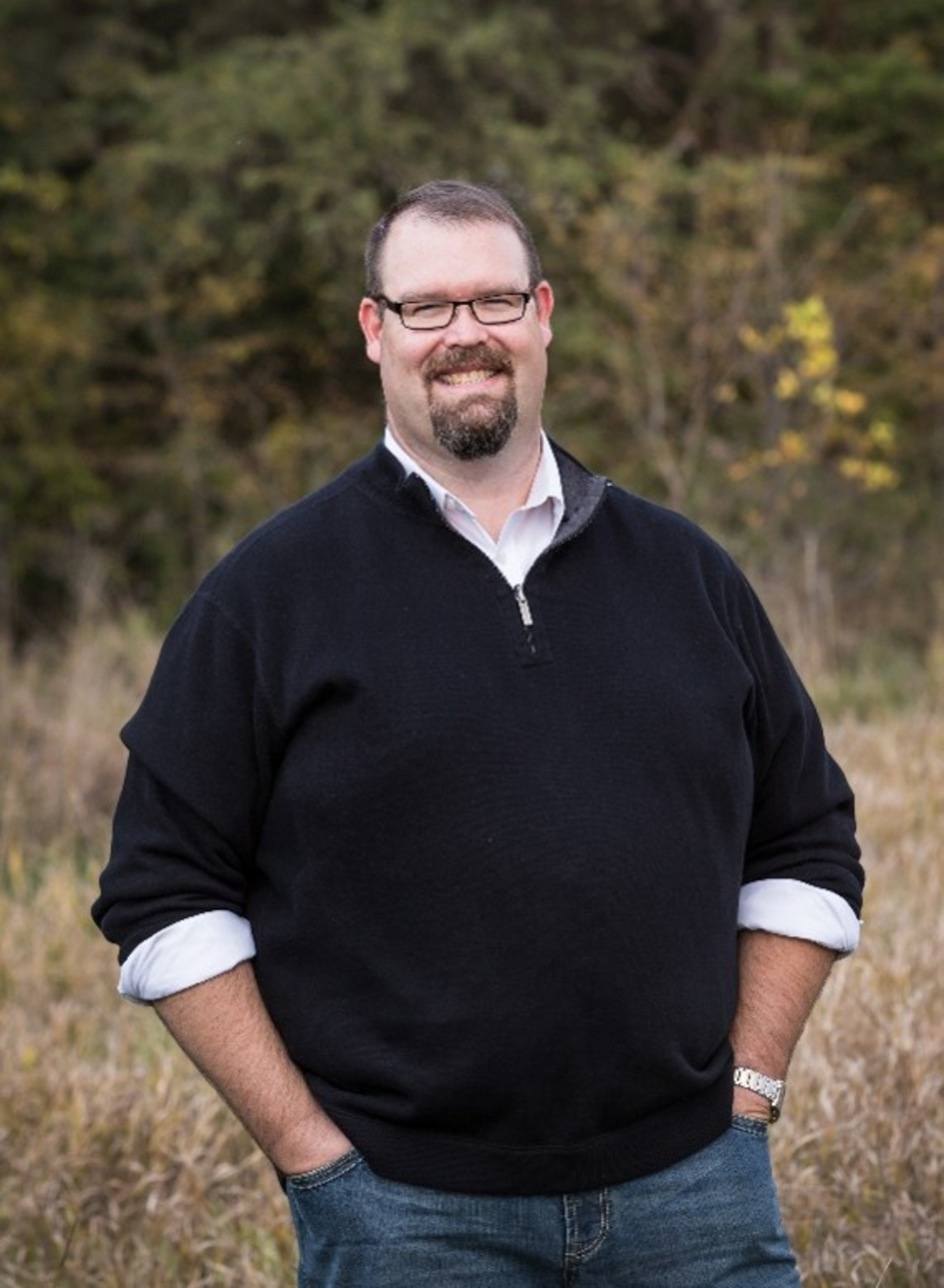 Smiling man wearing glasses and a dark sweater stands outdoors in a grassy area with trees.