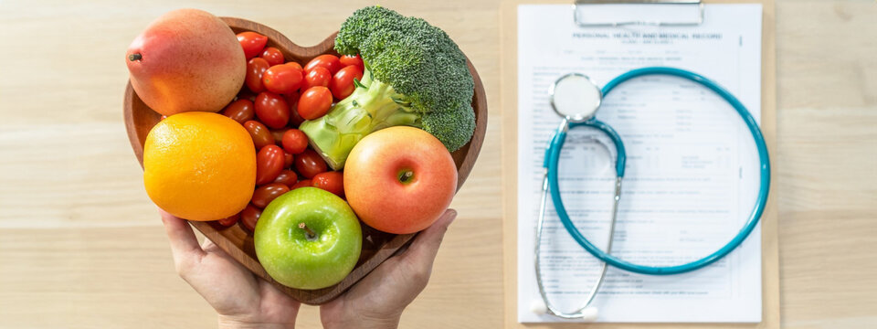 Heart shaped bowl of fruit being held over table with clipboard, chart, and stethoscope. 