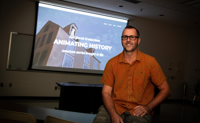 Michael Burton sits in a dark classroom in front of a large screen displaying the Animating History webpage. 