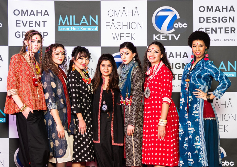 Seven individuals pose on a red carpet, wearing hand crafted looks that were showcased at Omaha Fashion Week. 